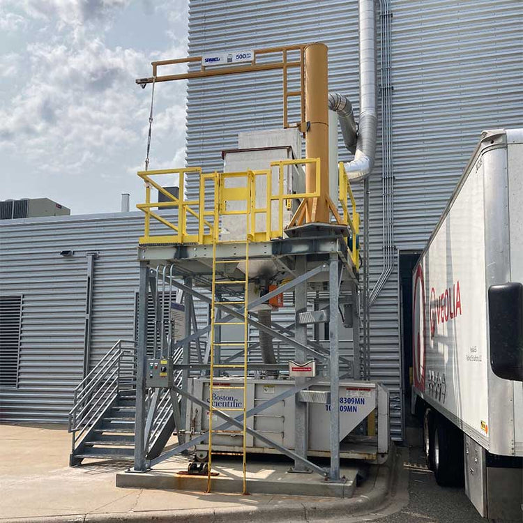 Industrial setup with a yellow platform and metal structure near a building, with a truck in the foreground.